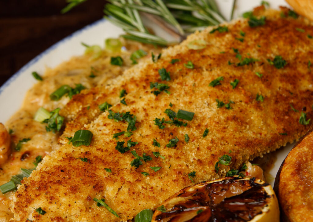 A breaded fish fillet at a Gulf seafood restaurant, garnished with green herbs, grilled lemon, and rosemary sprig, alongside a glass of white wine.